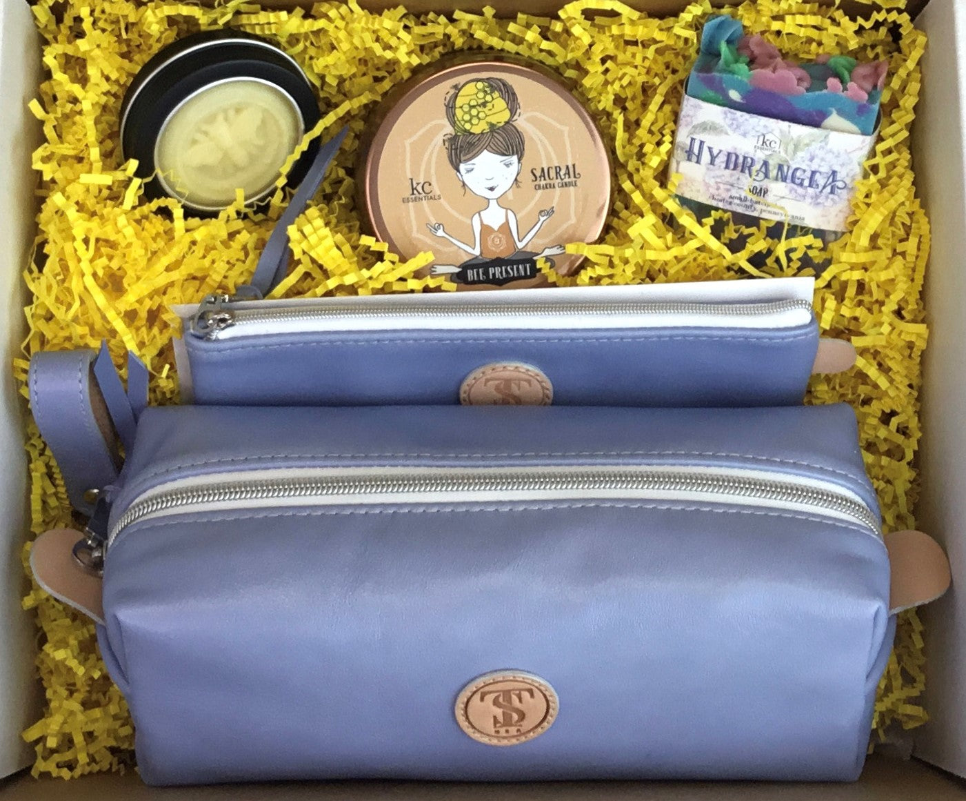 Town & Shore Handcrafted Serenity Gift set featuring T5 Leather bath kit and brush bag shown in light periwinkle blue calfskin leather. KC Essentials artisan made vegan bar soap, scented soy chakra candle in a bronze metal tin with a lady in a seated yoga pose on lid, and natural solid lotion bar in metal tin. Arranged in gift box with sustainable eco-friendly yellow crinkle paper.