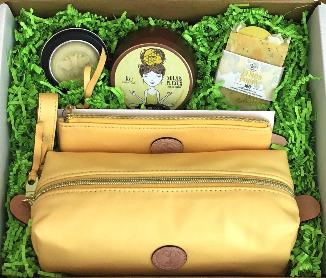 Town & Shore Handcrafted Serenity Gift set featuring T5 Leather bath kit and brush bag shown in saffron yellow calfskin leather. KC Essentials artisan made vegan bar soap, scented soy chakra candle in a bronze colored metal tin with a lady in a seated yoga pose on lid, and natural solid lotion bar in metal tin. Arranged in gift box with sustainable eco-friendly green crinkle paper.