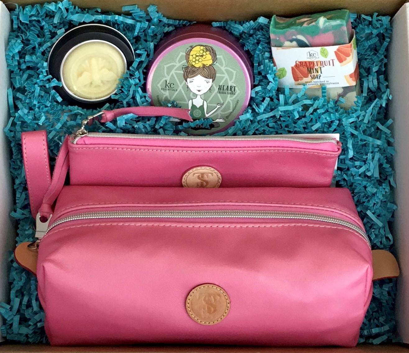Town & Shore Handcrafted Serenity Gift set featuring T5 Leather bath kit and brush bag shown in baby pink calfskin leather. KC Essentials artisan made vegan bar soap, scented soy chakra candle in a pink metal tin with a lady in a seated yoga pose on lid, and natural solid lotion bar in metal tin. Arranged in gift box with sustainable eco-friendly turquoise crinkle paper.