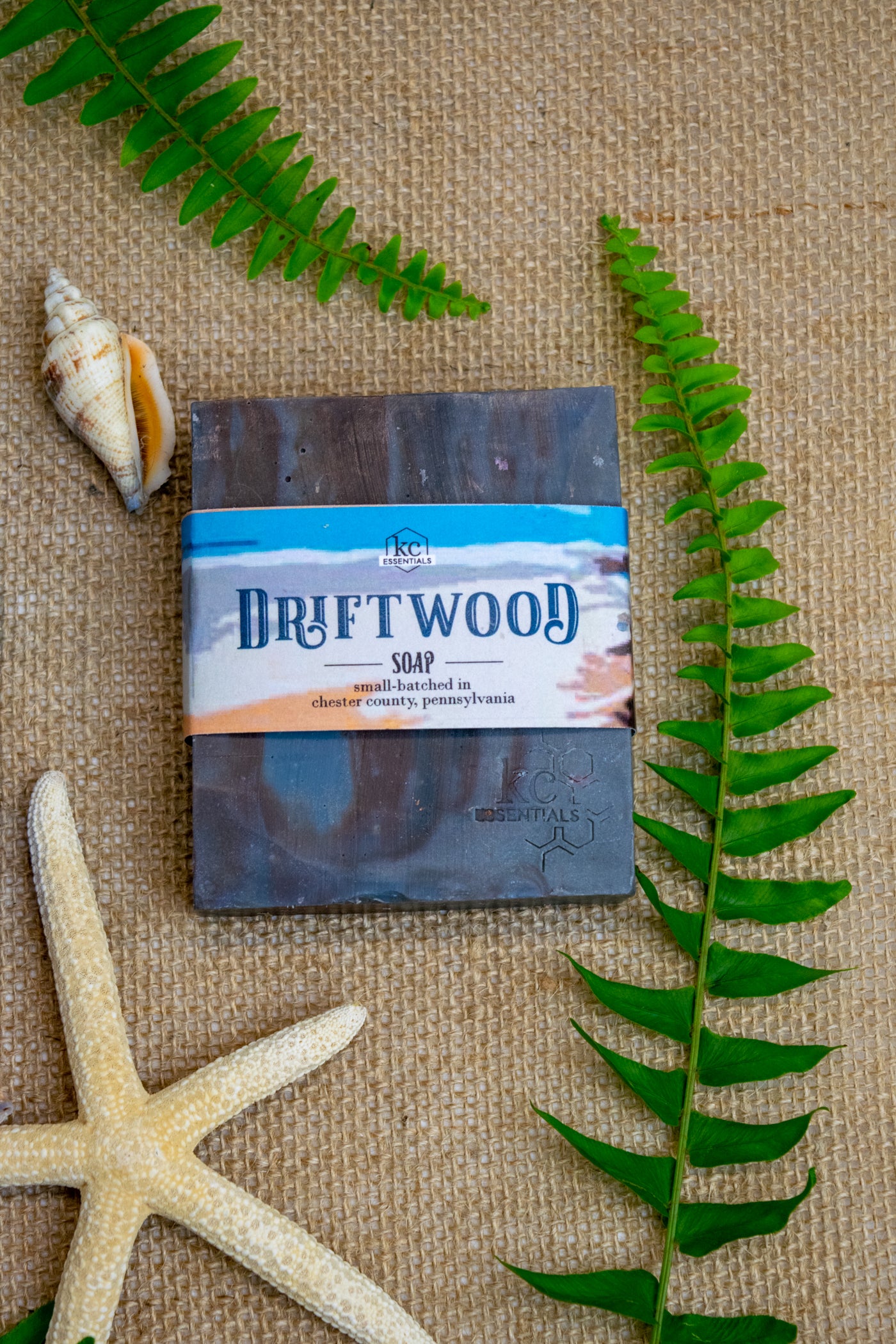 Streaks of brown and shades of grey form this artisanal soap bar on a backrop of burlap cloth with seashells and fern leaves.