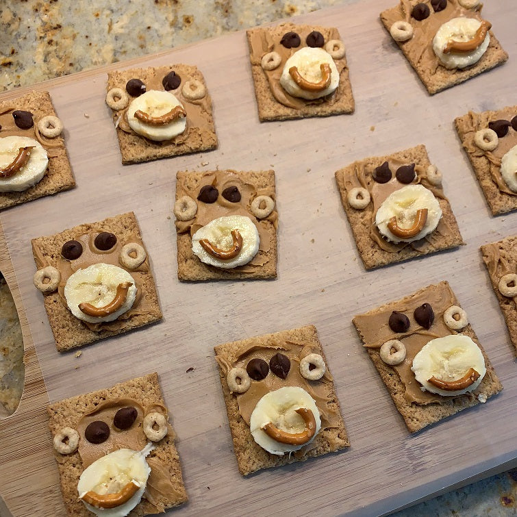 picture of graham crackers that look like monkey faces with peanut butter, chocalate chips, banana, cheerios and pretzal pieces.