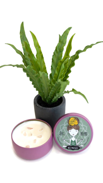 Small plant next to the Scented Heart Chakra Candle in shiny purple colored Lidded Tin with pink Rose Quartz crystals in the soy wax. Lid shows a girl in a seated yoga pose