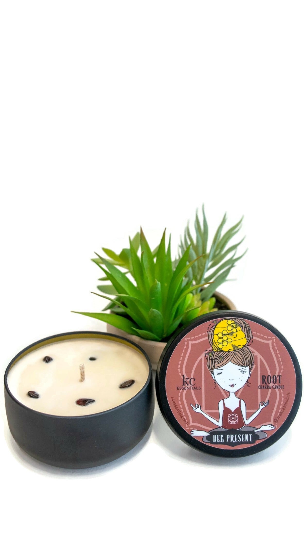 Small plant next to the Scented Root Chakra Candle in matt black  Lidded Tin with Garnet crystals in the soy wax. Lid shows a girl in a seated yoga pose