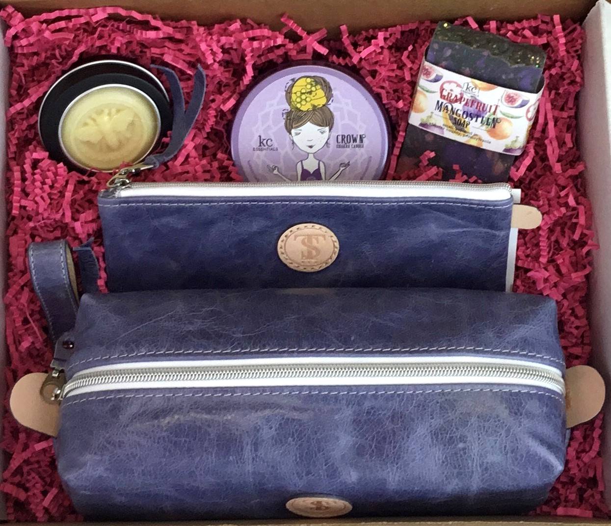 Town &amp; Shore Handcrafted Serenity Gift set featuring T5 Leather bath kit and brush bag shown in denim blue calfskin leather. KC Essentials artisan made vegan bar soap, scented soy chakra candle in a purple metal tin with a lady in a seated yoga pose on lid, and natural solid lotion bar in metal tin. Arranged in gift box with sustainable eco-friendly bright pink crinkle paper.