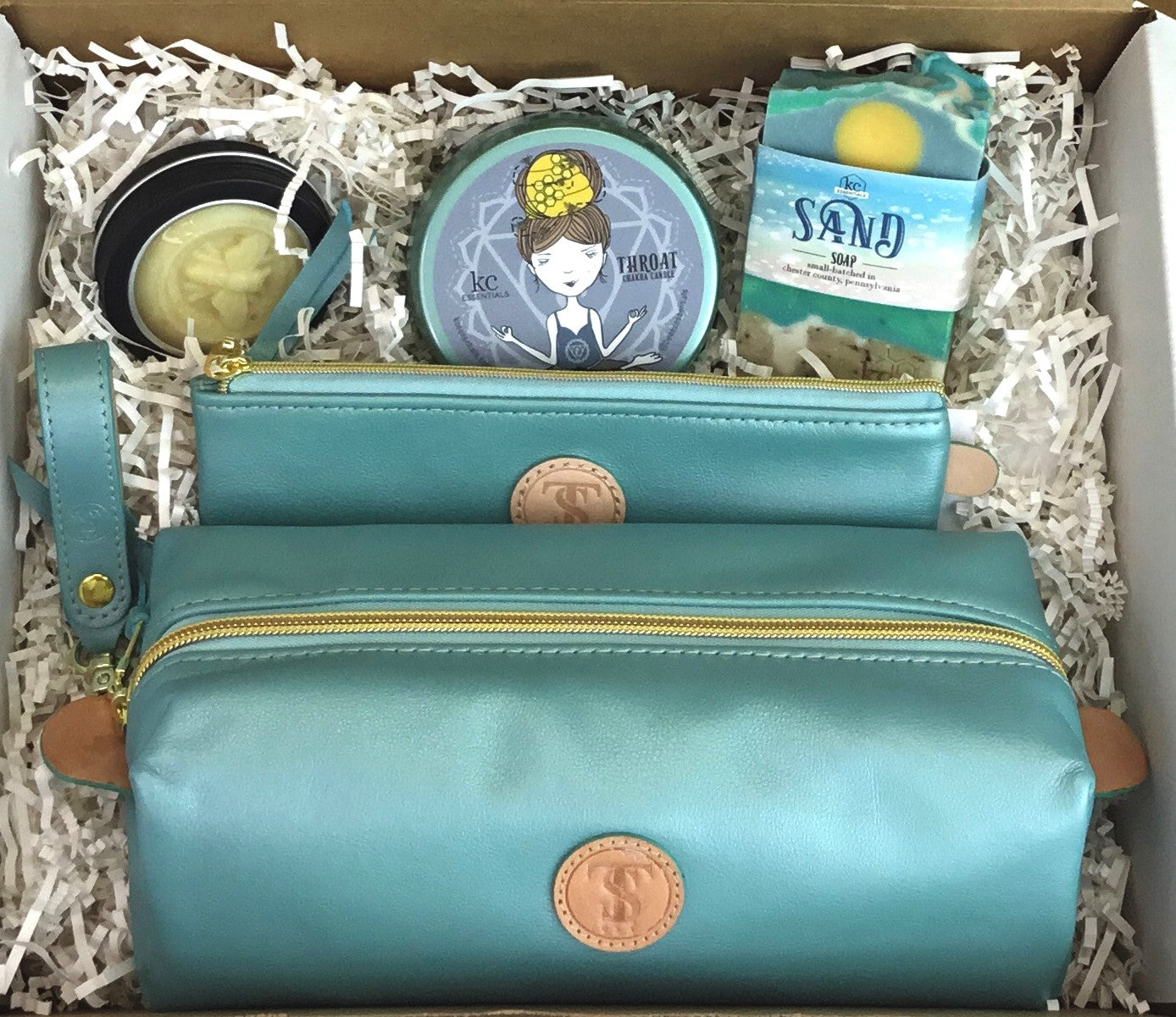 Town &amp; Shore Handcrafted Serenity Gift set featuring T5 Leather bath kit and brush bag shown in turqouise calfskin leather. KC Essentials artisan made vegan bar soap, scented soy chakra candle in a turqoise colored metal tin with a lady in a seated yoga pose on lid, and natural solid lotion bar in metal tin. Arranged in gift box with sustainable eco-friendly white crinkle paper.
