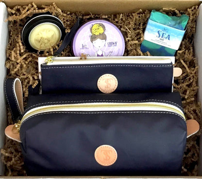 Town &amp; Shore Handcrafted Serenity Gift set featuring T5 Leather bath kit and brush bag shown in navy blue calfskin leather. KC Essentials artisan made vegan bar soap, scented soy chakra candle in a purple metal tin with a lady in a seated yoga pose on lid, and natural solid lotion bar in metal tin. Arranged in gift box with sustainable eco-friendly craft crinkle paper.