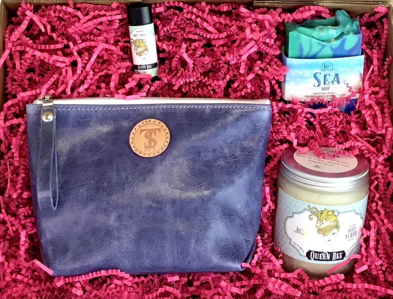 Town &amp; Shore Handcrafted Serenity Gift set featuring T5 Leather bath kit and brush bag shown in denim blue calfskin leather. KC Essentials artisan made vegan bar soap, essential oils body scrub and soothing lip balm. Arranged in gift box with sustainable eco-friendly bright pink crinkle paper.