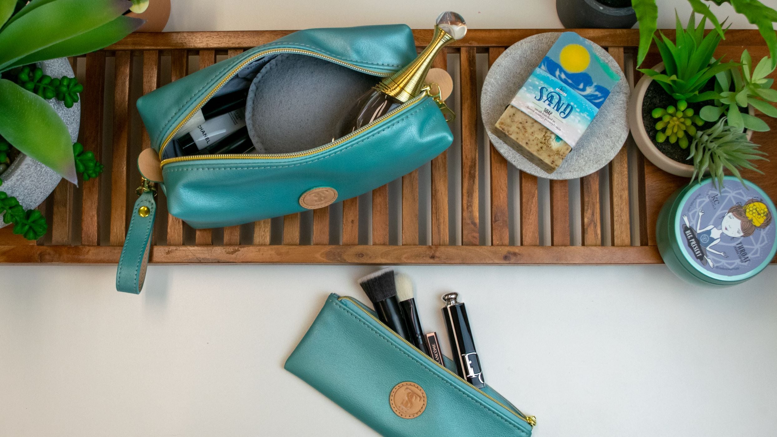 Town & Shore Handcrafted T5 Leather bath kit and brush bag shown in turquoise calfskin leather. Shown organizing cosmetics, toiletries, brushes. Shown on teak wood platform surrounded by tropical plants with Artisan made vegan bar soap and scented soy chakra candle in a metal tin.
