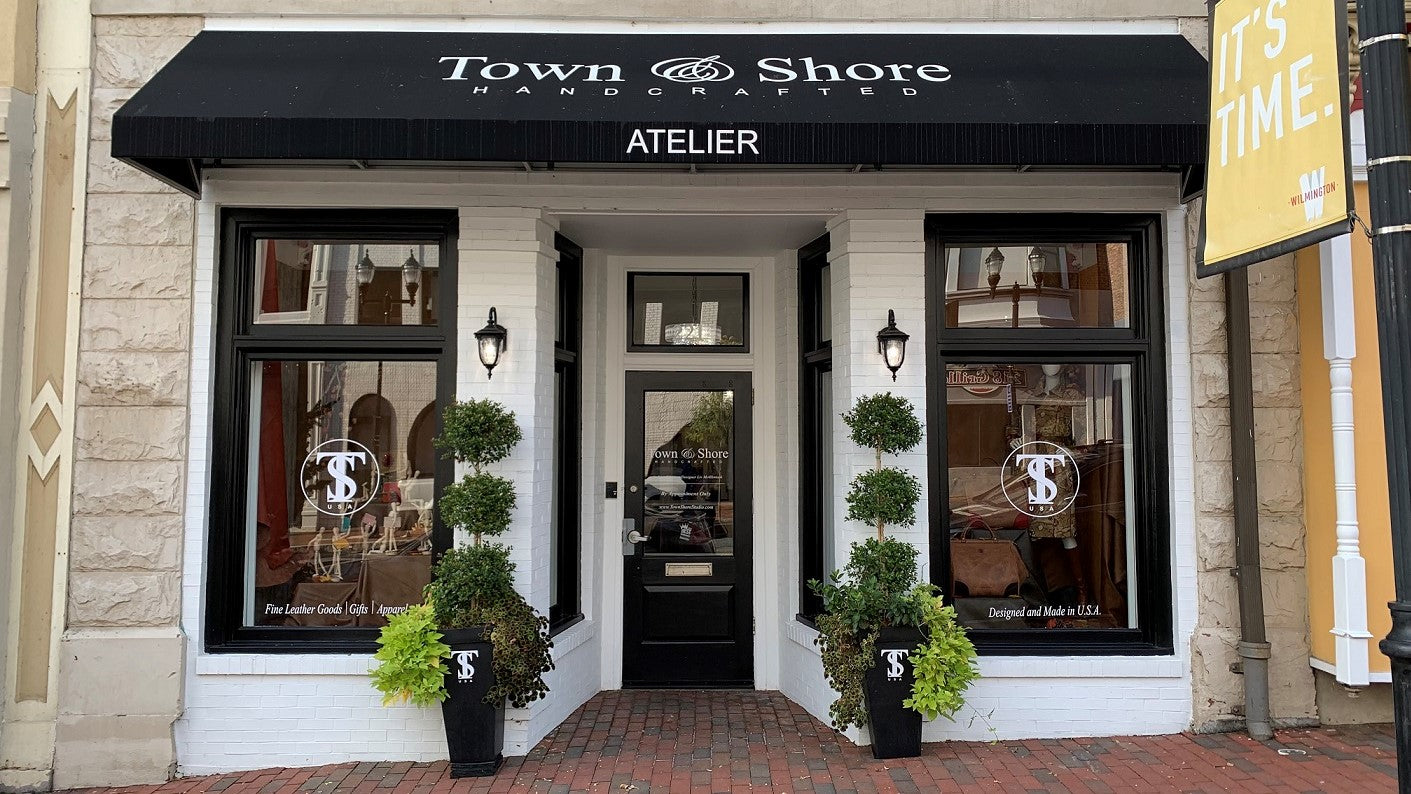 Entrance of the Town & Shore Handcrafted Flagship Store front. White Building with Windows trimmed in black and Large Black signage awinging with shop name. Tall black planters with green topiaries and hanging plants grace the doorway.